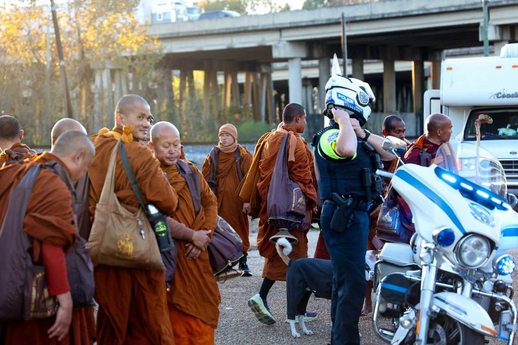 A line of monks in orange robes prepare to walk down a road with a police escort