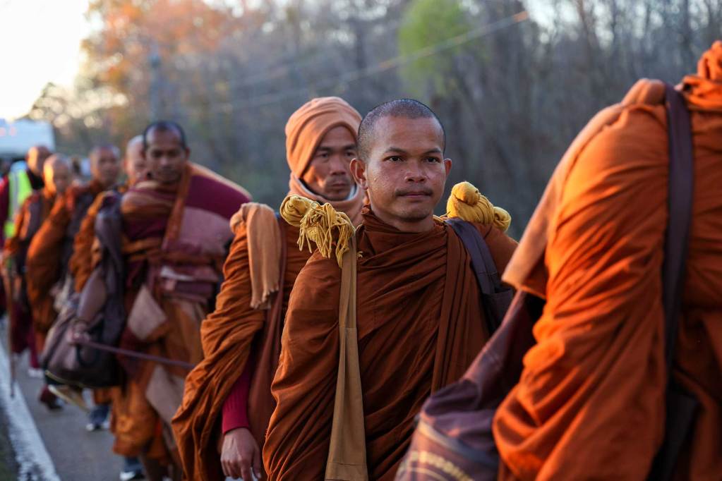 A line of monks in orange robes walk down a paved road in Mississippi