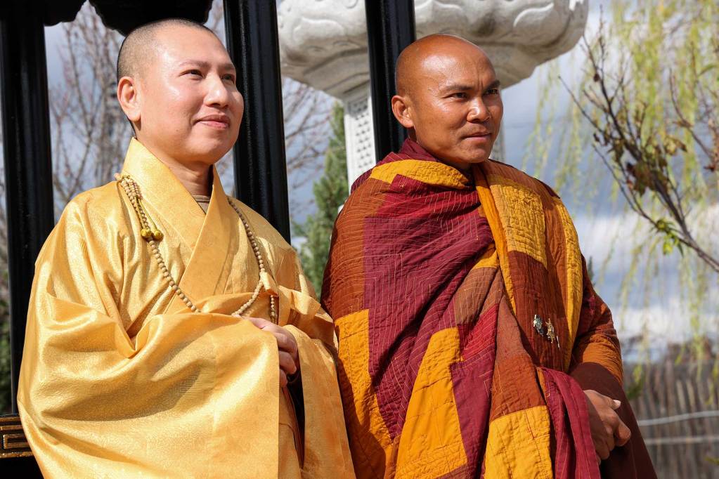 Two monks standing outside, one in golden robes and one in orange patchwork robes