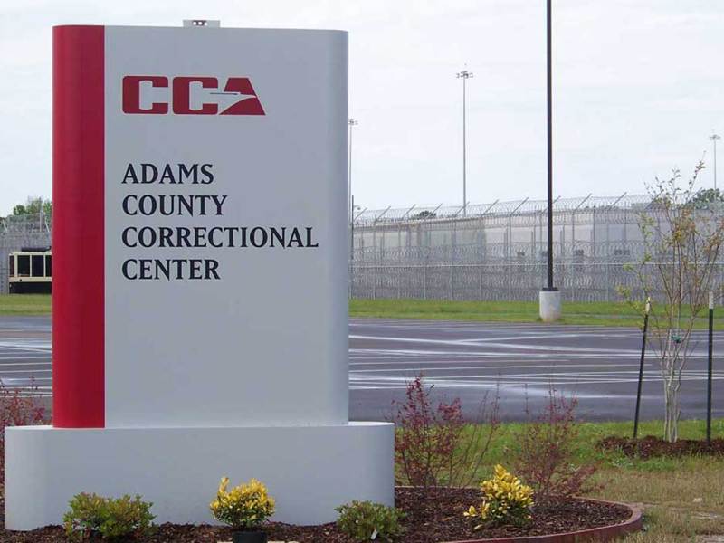The sign for Adams County Correctional Center, barbed wire fencing visible around the facility behind it