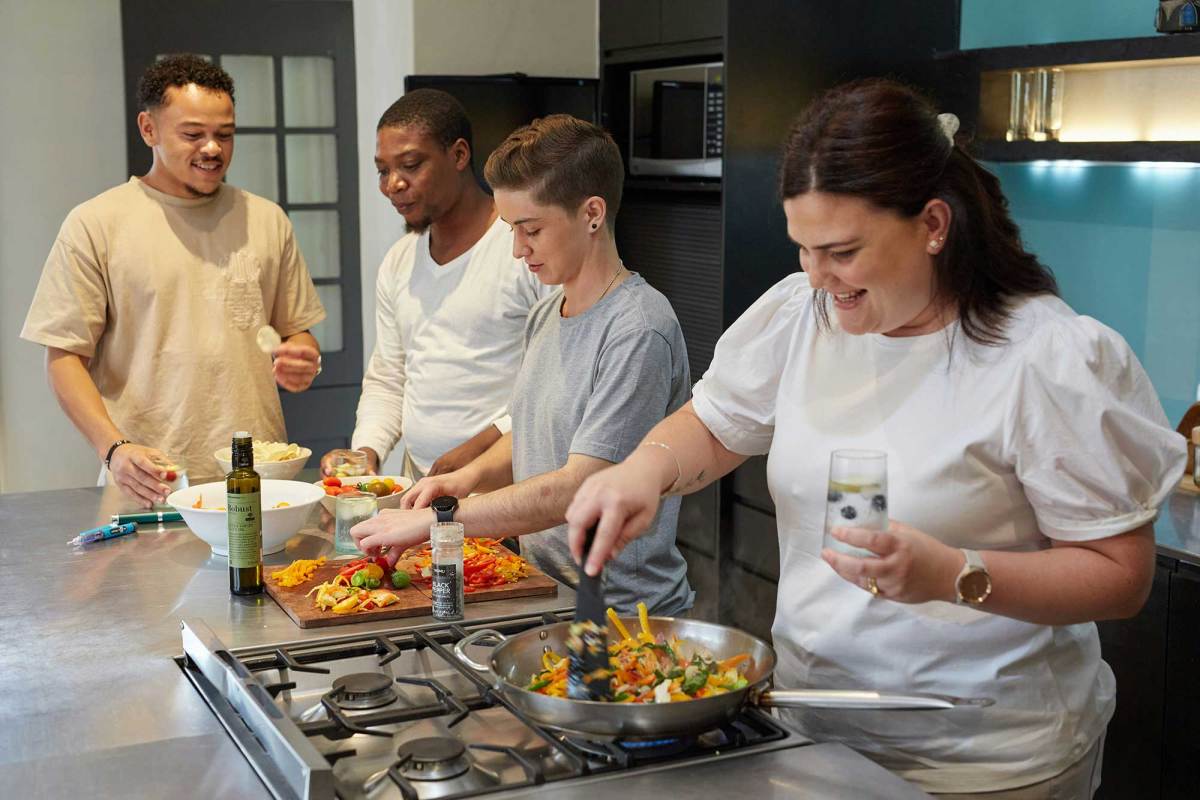 Two men and two women lined up at a kitchen counter, with the woman closest to the camera stir-frying vegetables on a stovetop.