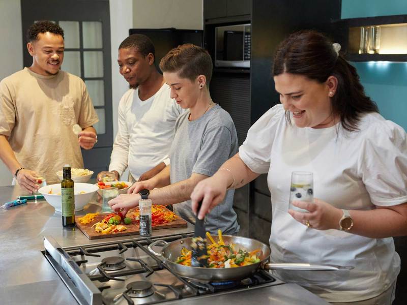 Two men and two women lined up at a kitchen counter, with the woman closest to the camera stir-frying vegetables on a stovetop.