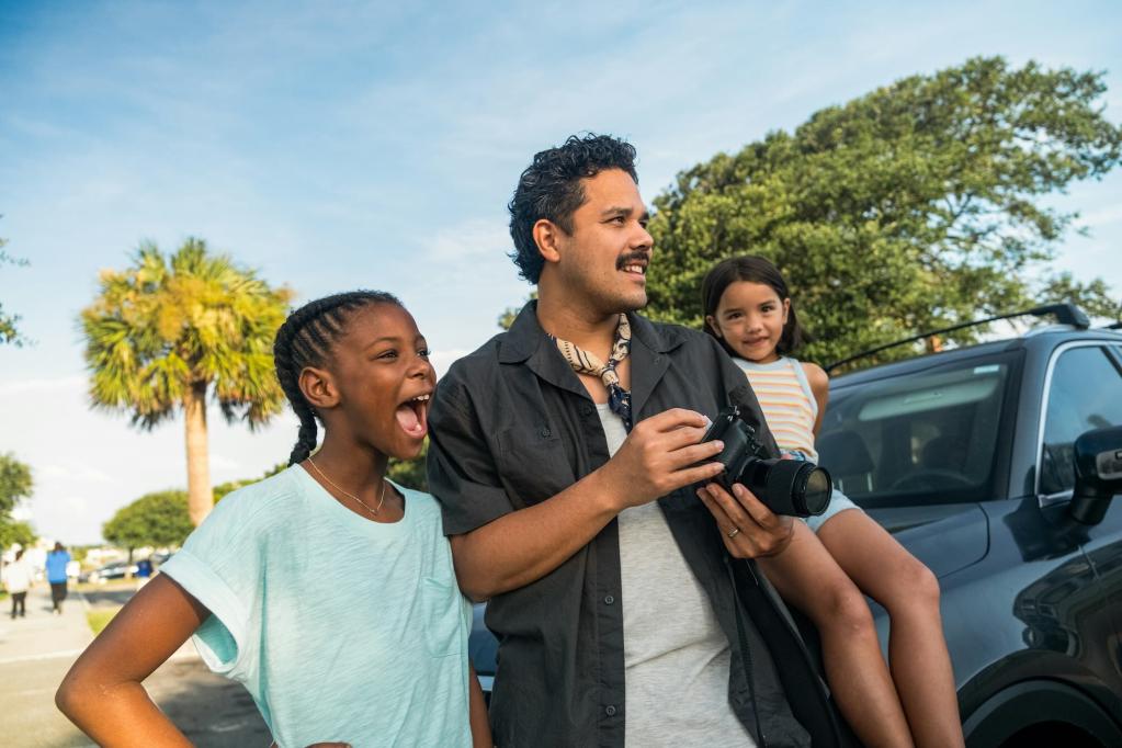 Two girls and a man looking off to the side. The man is holding a camera. One of the girls is seated on the hood of a truck.