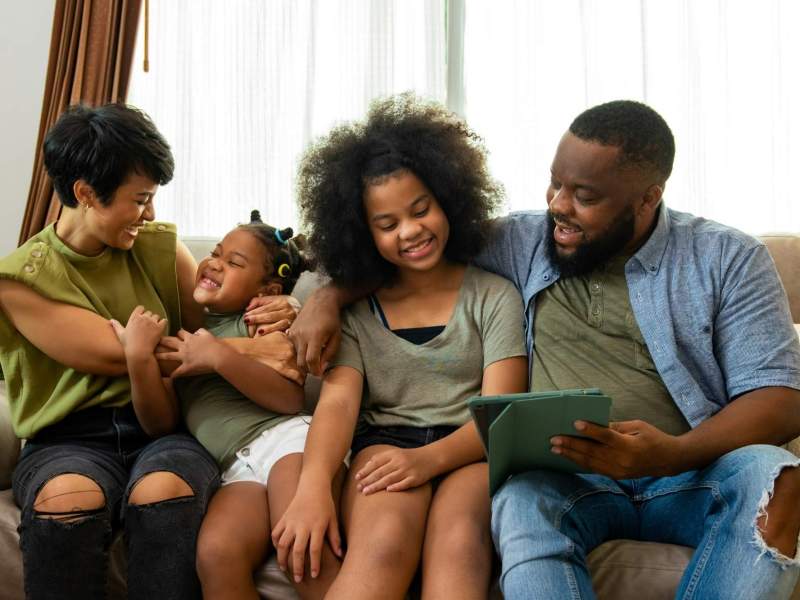 A woman, man and two girls seated on a couch. The man and one of the girls are looking at a tablet computer.