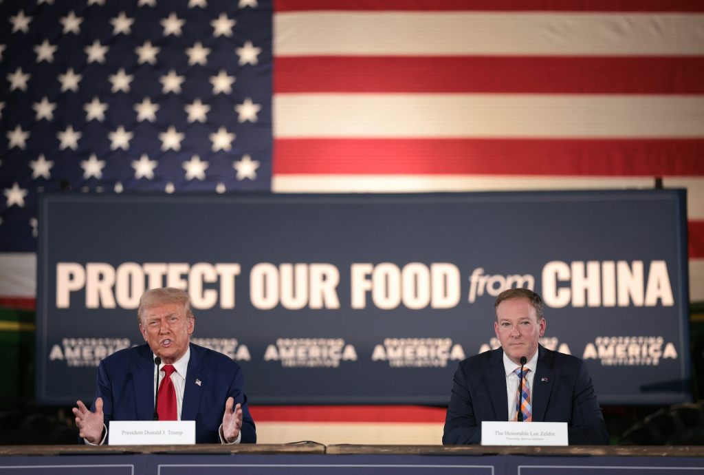 Donald Trump and Lee Zeldin seated at a desk in front of a sign that reads ‘Protect our food from China’