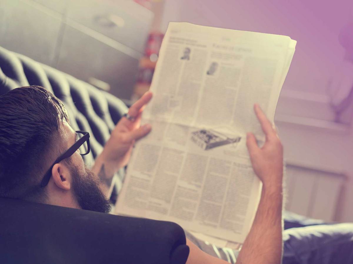 A man laying on a black couch in a home reading the newspaper