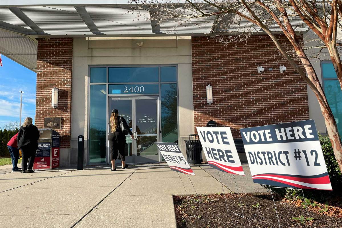 Several “Vote Here” yard signs outside a brick building. A woman is about to enter the building through its glass door.