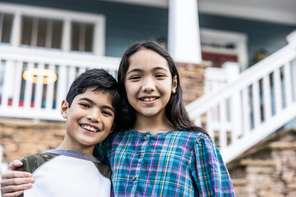 A young boy and older girl stand together smiling in front of a house.