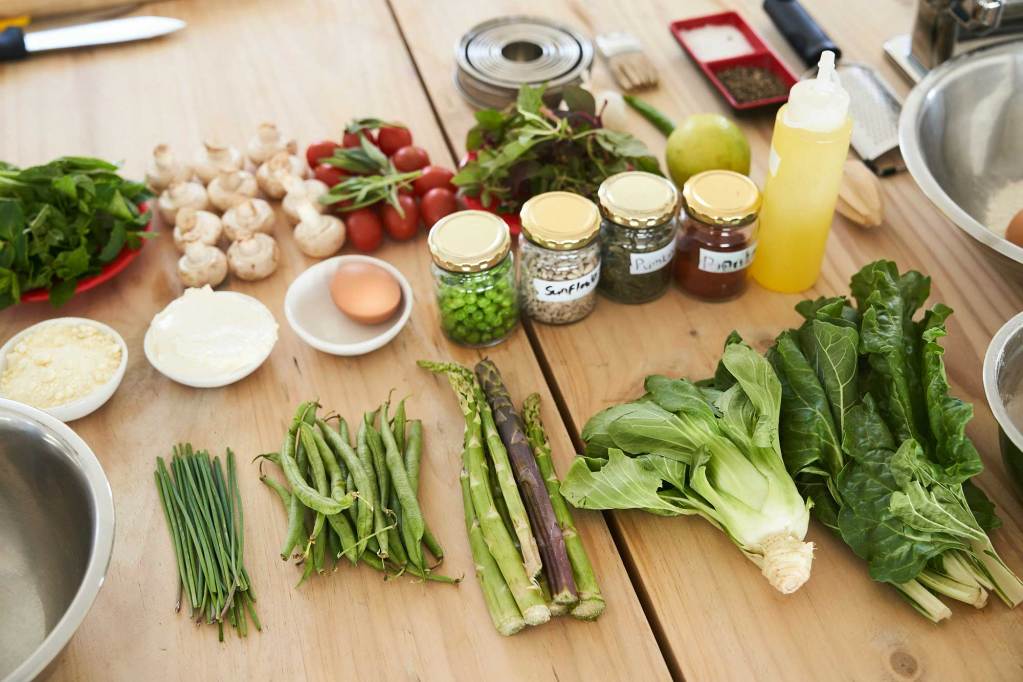 A counter with numerous vegetables lined up including beans, asparagus, leaks, lettuce, tomatoes and onions.