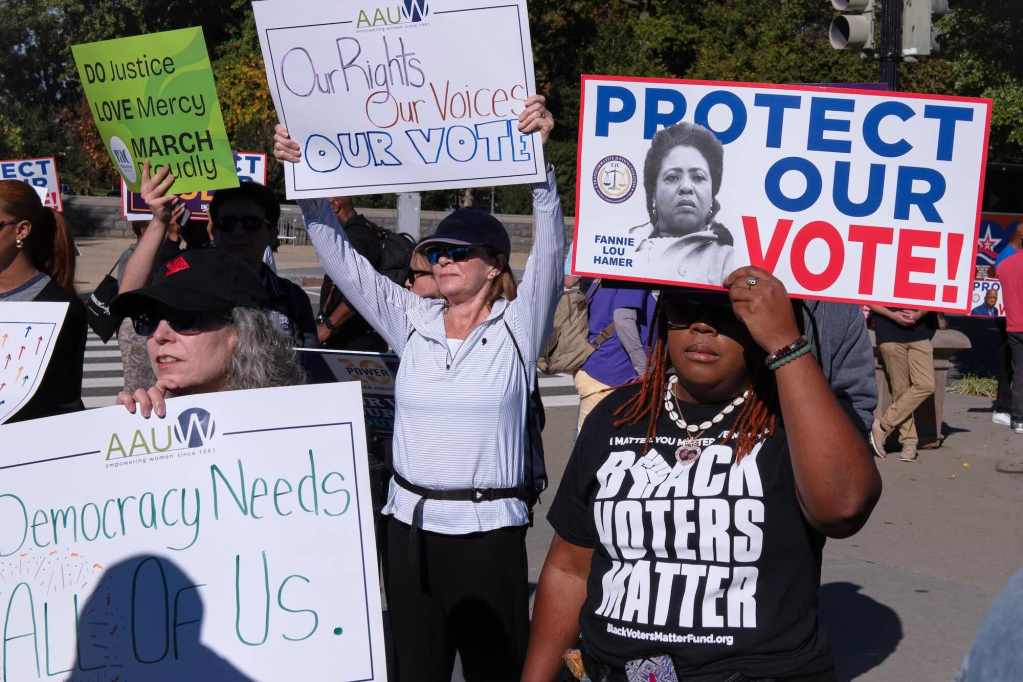 people stand around holding protest signs supporting voting rights. One woman wearing a black tshirt that says "BLACK VOTERS MATTER" is holding up a sign with a black and white photo of Fannie Lou Hamer with the words "PROTECT OUR VOTE"