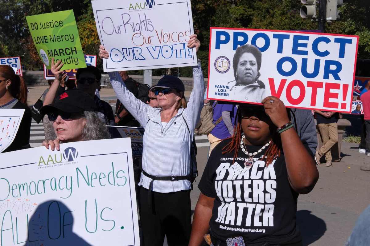 people stand around holding protest signs supporting voting rights. One woman wearing a black tshirt that says "BLACK VOTERS MATTER" is holding up a sign with a black and white photo of Fannie Lou Hamer with the words "PROTECT OUR VOTE"