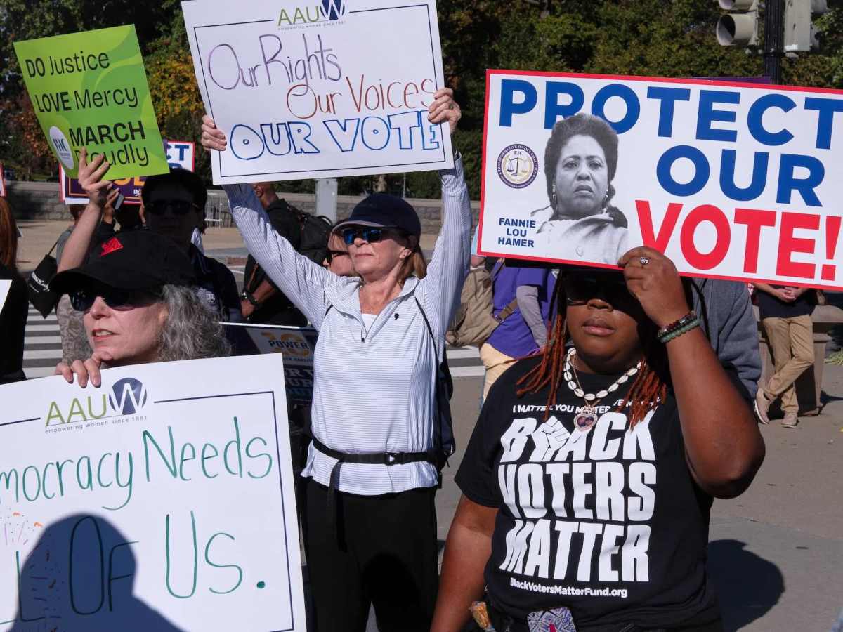 people stand around holding protest signs supporting voting rights. One woman wearing a black tshirt that says "BLACK VOTERS MATTER" is holding up a sign with a black and white photo of Fannie Lou Hamer with the words "PROTECT OUR VOTE"