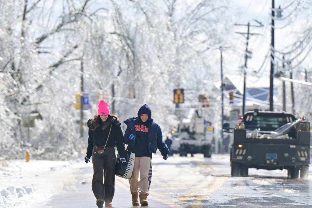 Two people walk down icy roads as utility trucks drive past