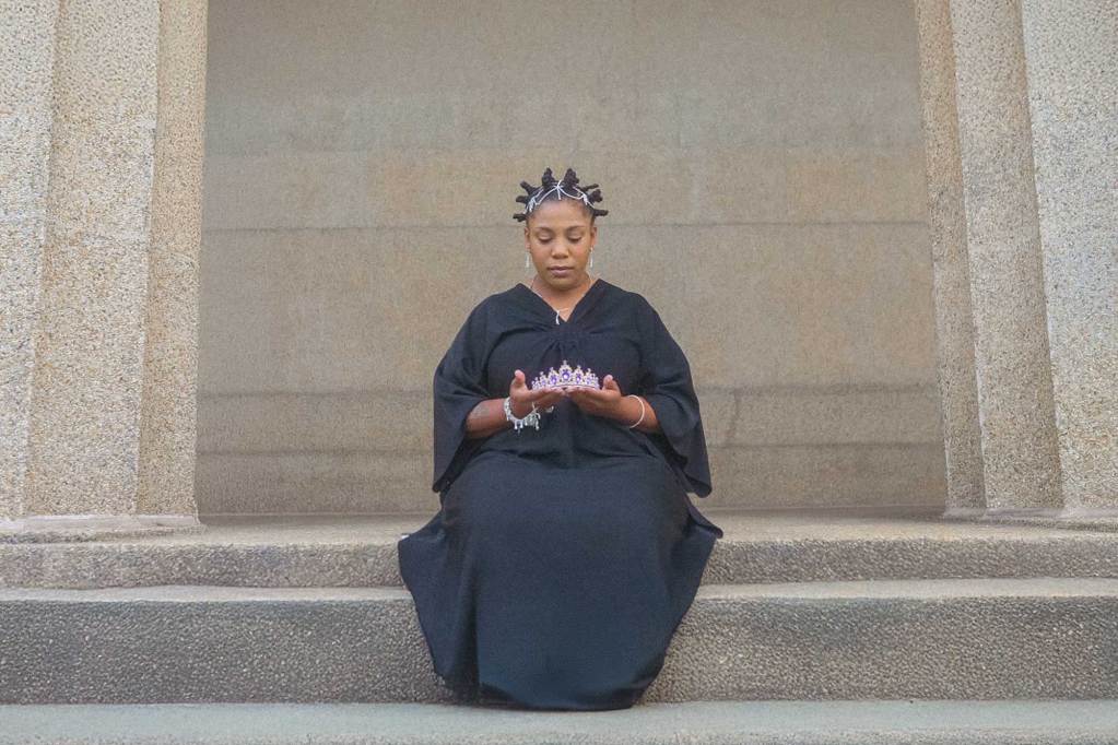 A woman in a black dress, sitting on a stairway outside and holding a crown