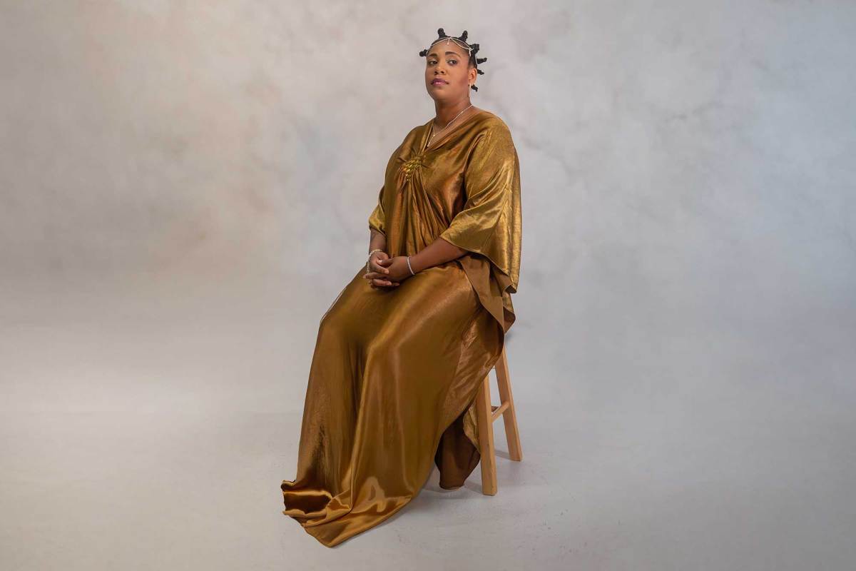 A woman in a long gold dress, sitting on a stool in a studio