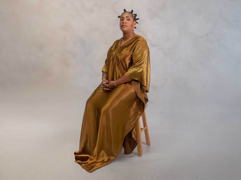 A woman in a long gold dress, sitting on a stool in a studio