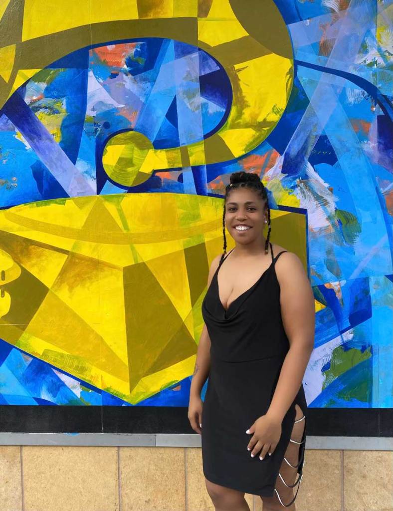 A woman in a black dress stands in front of a large piece of artwork depicting a Grammy award