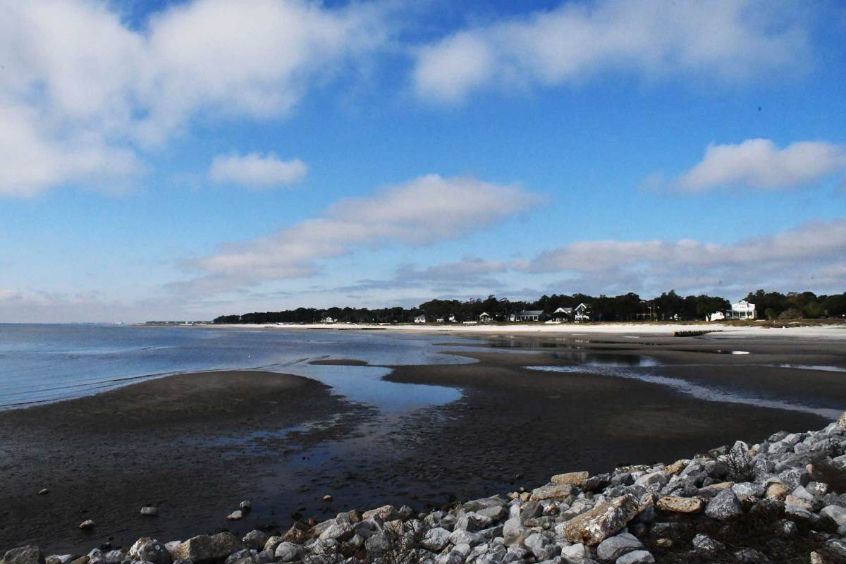 A wide coastal shoreline at low tide with patches of wet sand and shallow water, cloudy blue sky above, and houses lining the distant tree-covered shore.