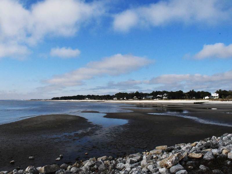 A wide coastal shoreline at low tide with patches of wet sand and shallow water, cloudy blue sky above, and houses lining the distant tree-covered shore.