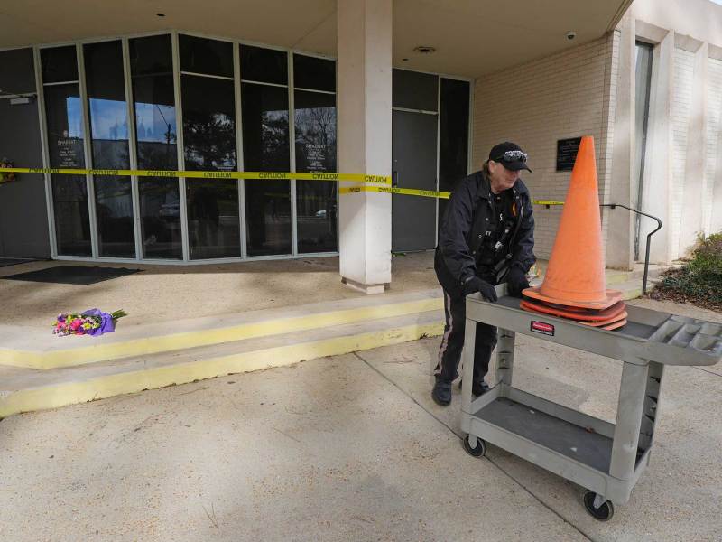 A person wheels orange cones past the entrance to Beth Israel Congregation, which is wrapped in yellow caution tape. A bouquet of flowers rests on the steps.