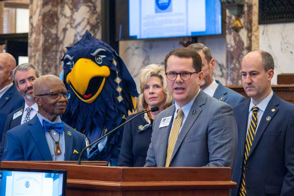 A men speaks at a podium. An eagle mascot looks on in the crowd behind him