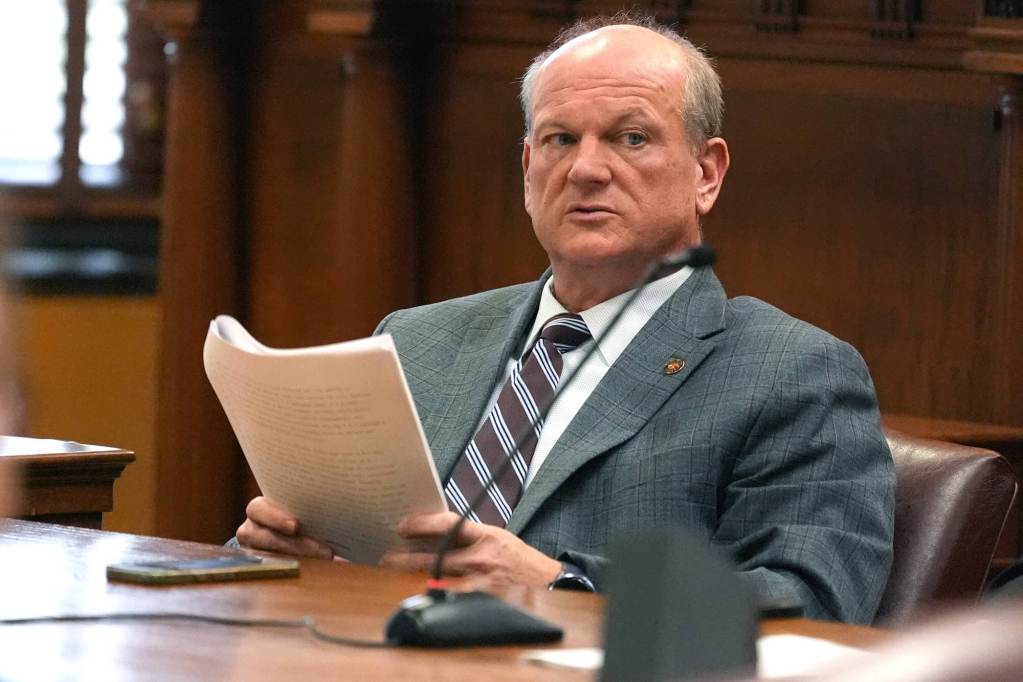 Brice Wiggins sits in a leather chair at a wooden table holding a paper copy of a bill.