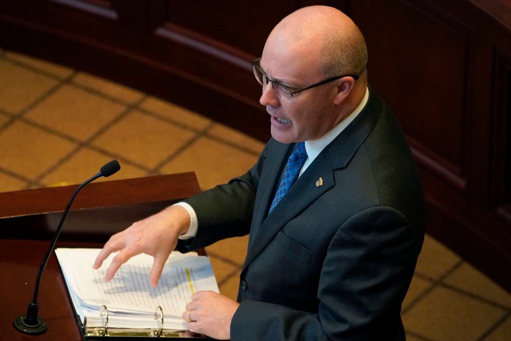 A man in a dark suit and blue tie speaking at a podium