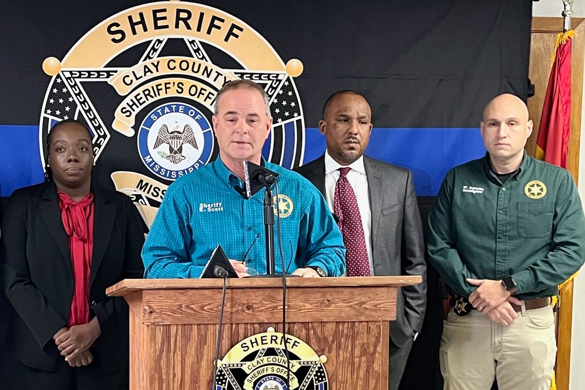 A woman and three men standing in a row behind a podium. One of the men, wearing a blue shirt, is speaking.