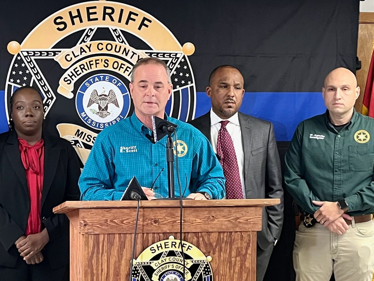 A woman and three men standing in a row behind a podium. One of the men, wearing a blue shirt, is speaking.