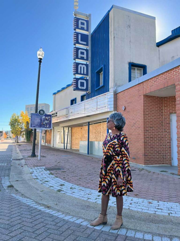 A woman in a colorful dress stands outside of a boarded up Alamo Theater