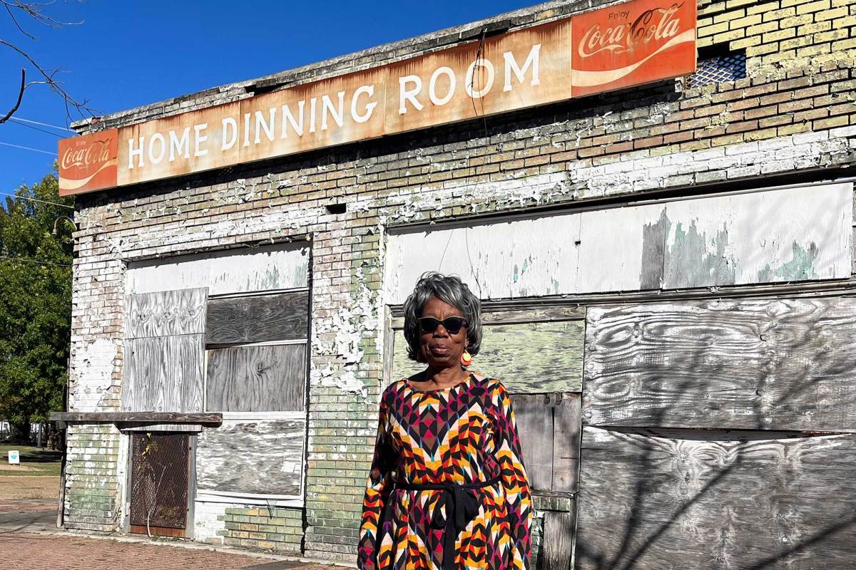 A woman in a colorful dress stands outside of a boarded up business with a sign that reads 'Home Dining Room'