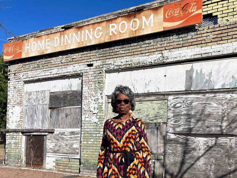 A woman in a colorful dress stands outside of a boarded up business with a sign that reads 'Home Dining Room'