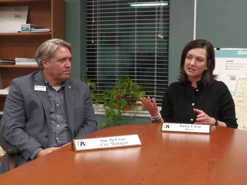 A man in a grey suit and a woman in a black shirt seated at a table and speaking