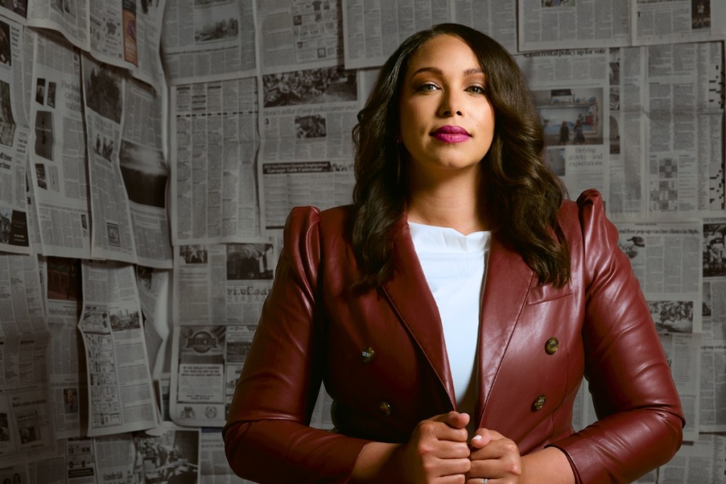 Georgia Ford standing confidently in a red blazer in front of a wall covered in newspapers