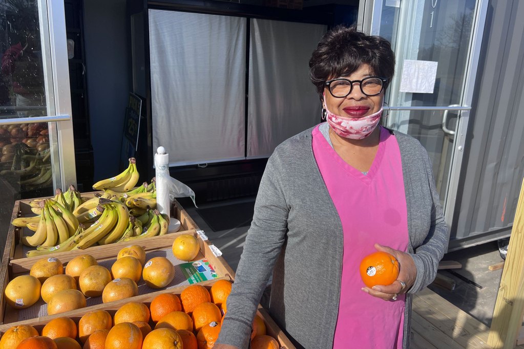 A woman holds an orange beside a fruit stand