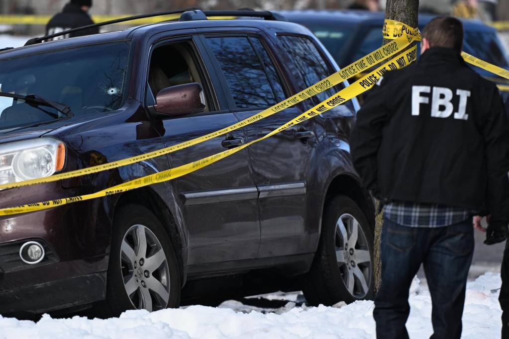 man wearing an FBI jacket approaches a vehicle. A bullet hole is in its windshield and is blocked by barricade tape.