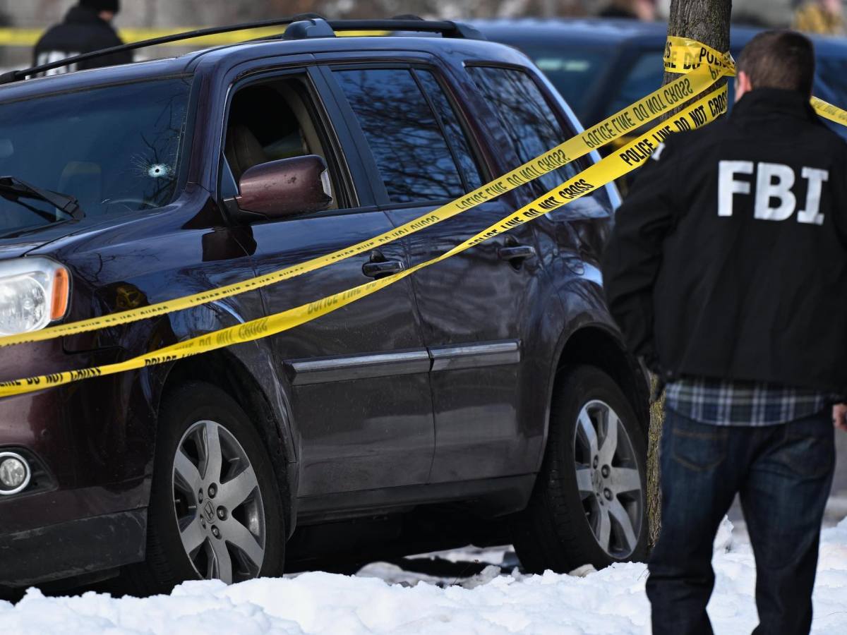 man wearing an FBI jacket approaches a vehicle. A bullet hole is in its windshield and is blocked by barricade tape.
