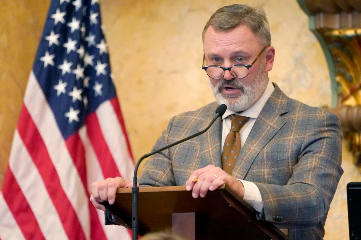 Jason White in a brown patterned suit speaking at a podium with the US flag visible behind him