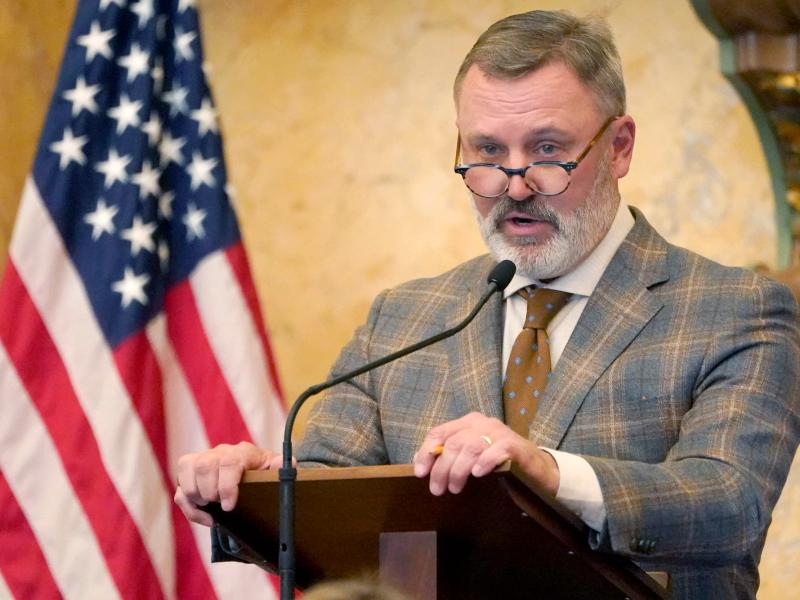 Jason White in a brown patterned suit speaking at a podium with the US flag visible behind him