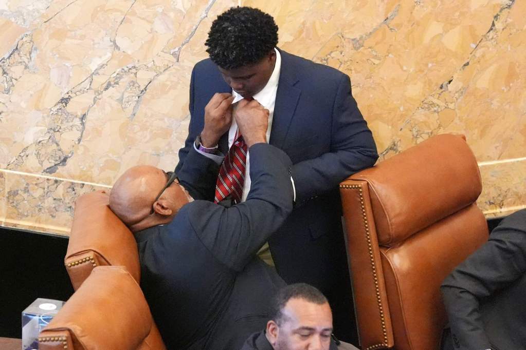 A seated older man helps a younger man with his suit tie