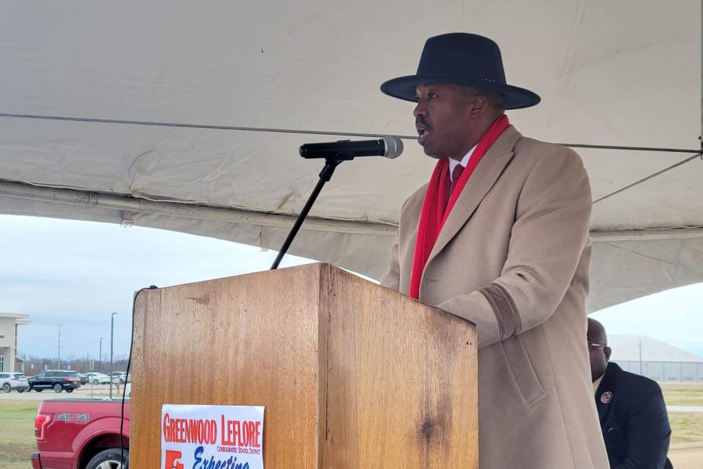 A man in a long coat speaks at a podium outside under a tent