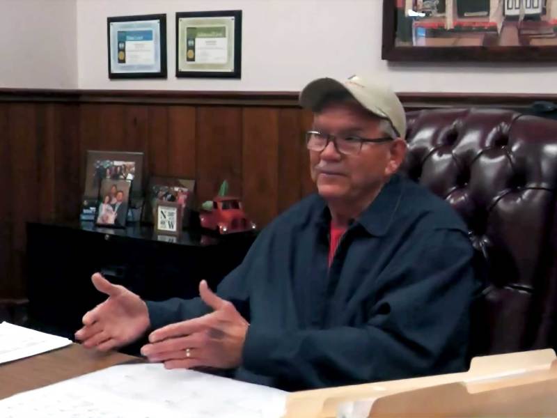 A man in baseball cap and glasses sits behind his office desk and speaks to an unseen interviewer