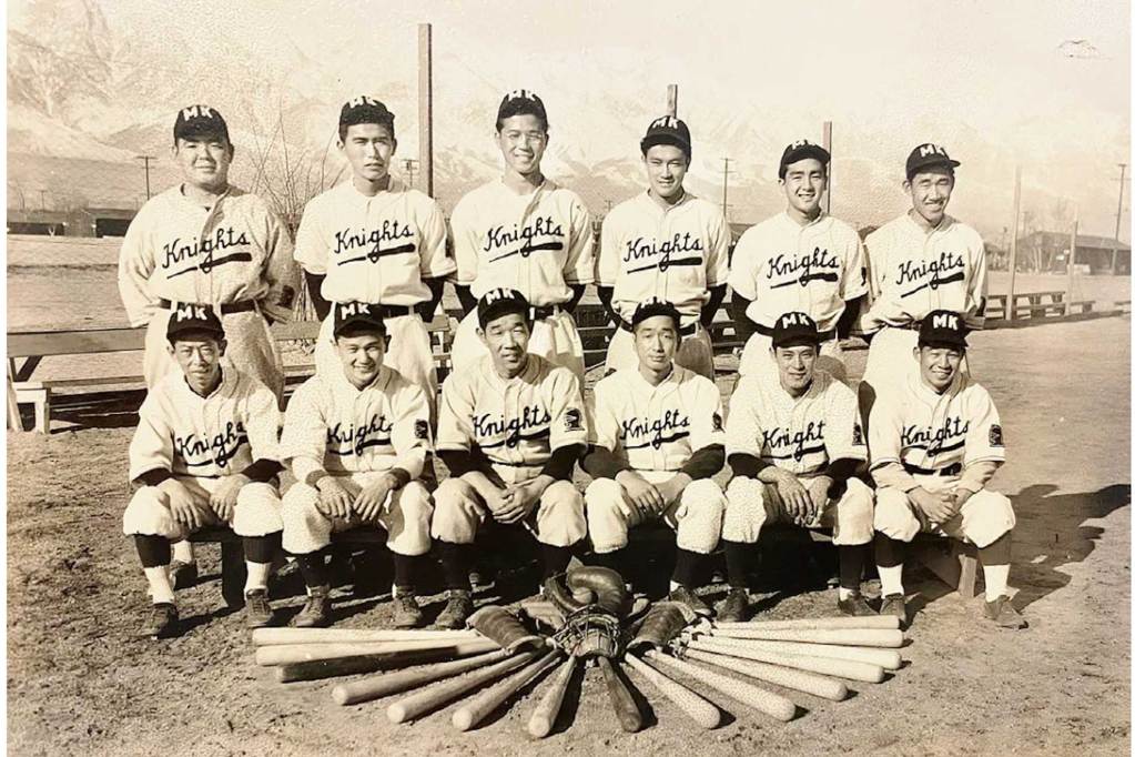 A black-and-white photo of two rows of men posing for a photo. All are wearing baseball uniforms, with the first row of men seated. A display of bats and gloves lay in front of them.