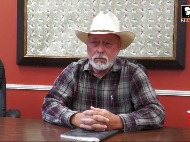 A man in a white cowboy hat sits at a conference table in a red walled room