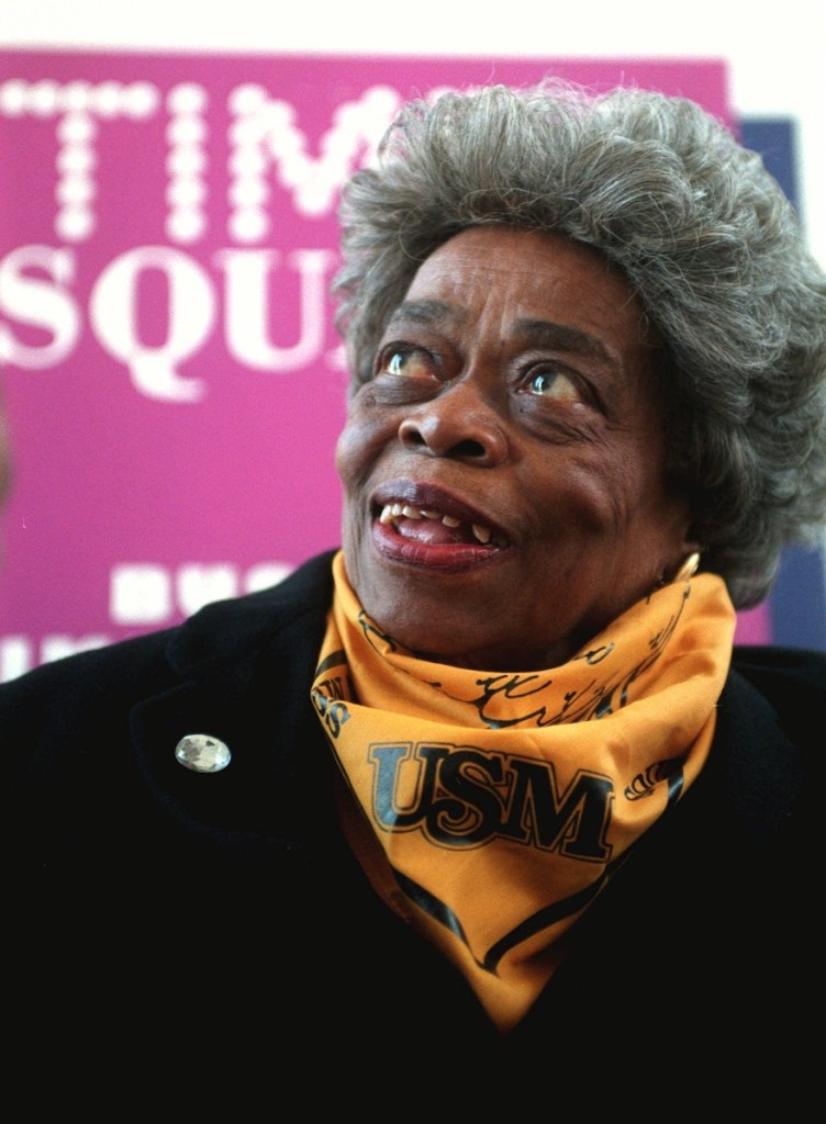 An older woman wearing a gold USM neck scarf smiles in front of a Times Square sign