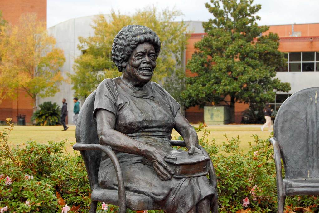 A bronze statue of a seated woman holding a book in her lap, situated in a green space beside large buildings