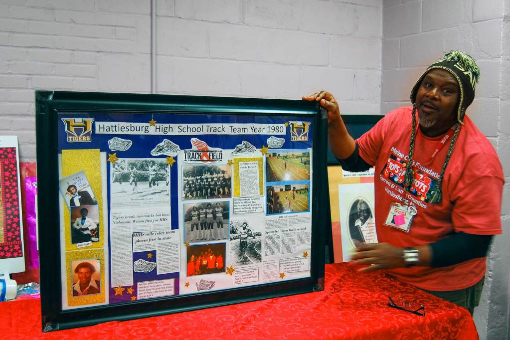A man in a red shirt stands beside a framed piece titled Hattiesburg High School Track Team Year 1980