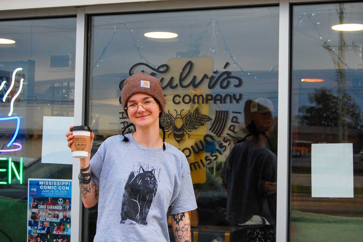 A woman stands outside of a shop holding a cup of coffee aloft