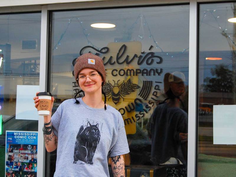 A woman stands outside of a shop holding a cup of coffee aloft