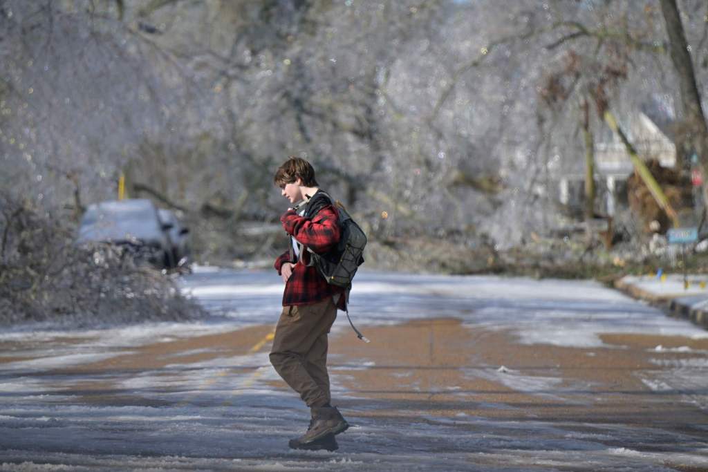 A man with a backpack walks across an icy road. Fallen trees are seen in the background